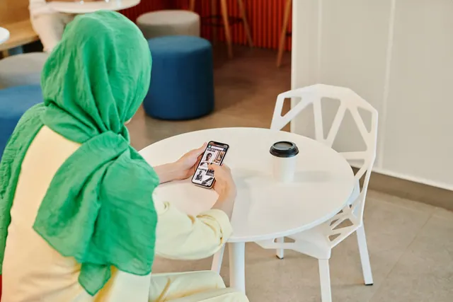 A woman wearing a green hijab is scrolling on her smartphone while sitting at a cafe table. Digital community models compared by purpose and governance