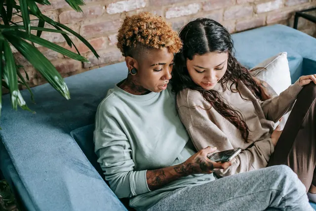 Concentrated young African American female scrolling feed in social network while resting on couch with interested Hispanic female. What goes wrong with parasocial relationships